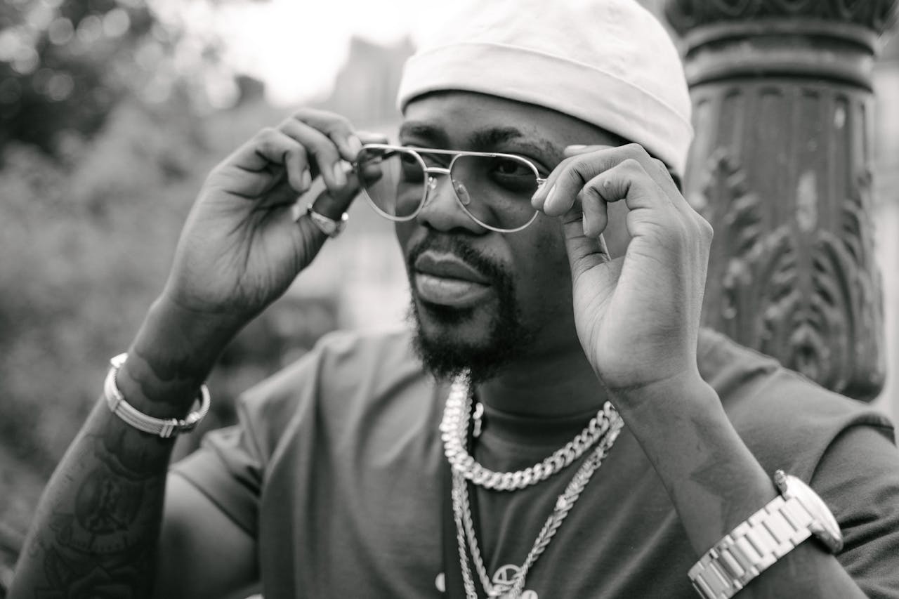 awards-img A fashionable man wearing sunglasses and jewelry, adjusting his glasses in an outdoor setting.
